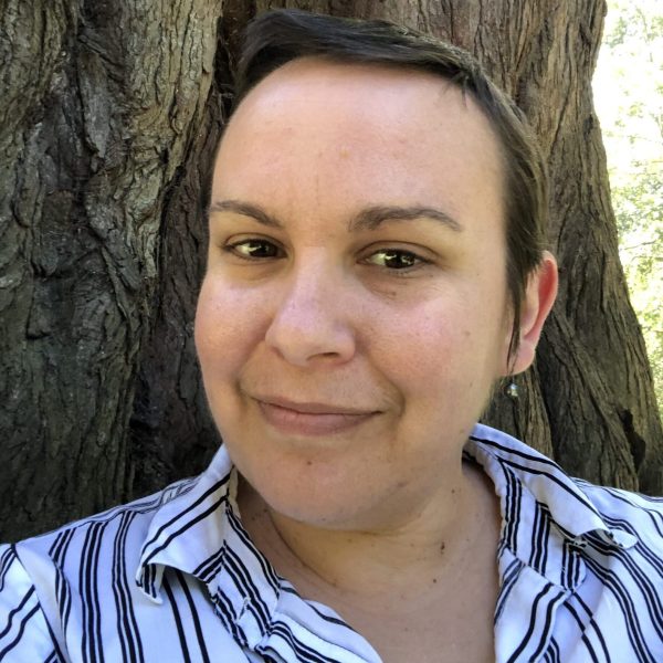 A person with short hair smiling gently, wearing a white shirt with black stripes, standing in front of a large tree with textured bark. The image is taken outdoors, with soft sunlight filtering through the background.